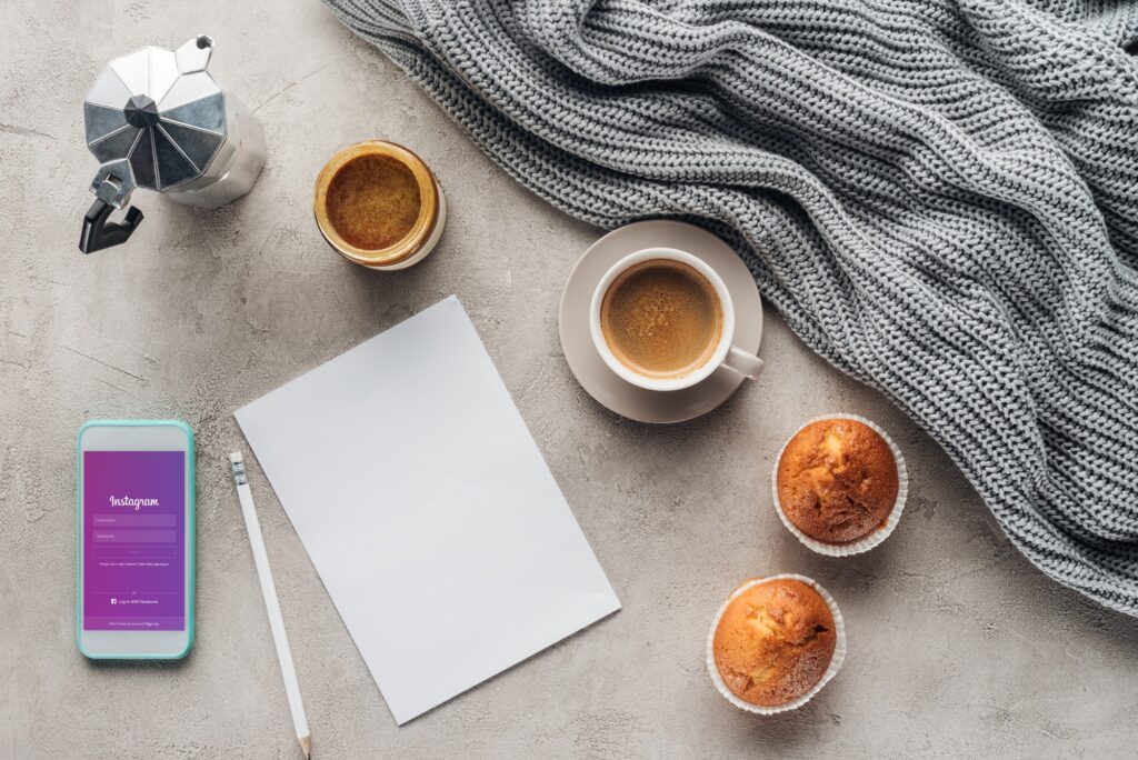 top view of cup of coffee with muffins, blank paper and smartphone with instagram app on screen on